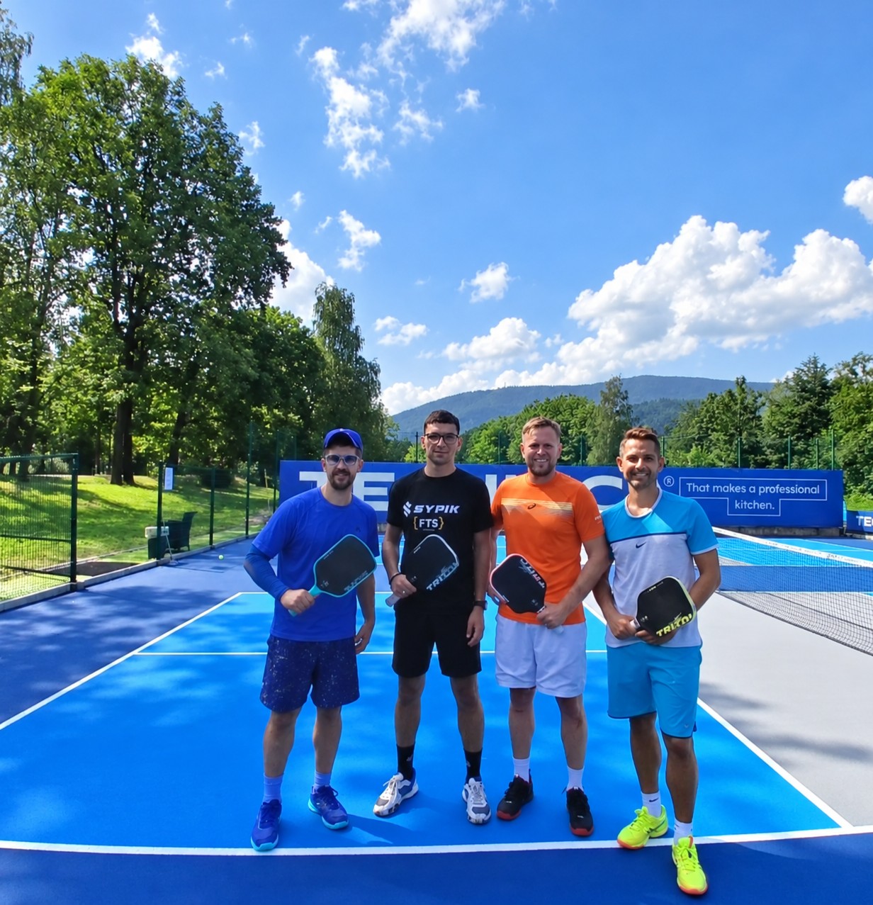 Czterech graczy na niebieskich kortach pickleballa w Klubie Tenisowym Kubala Ustroń, w otoczeniu zieleni i Beskidów